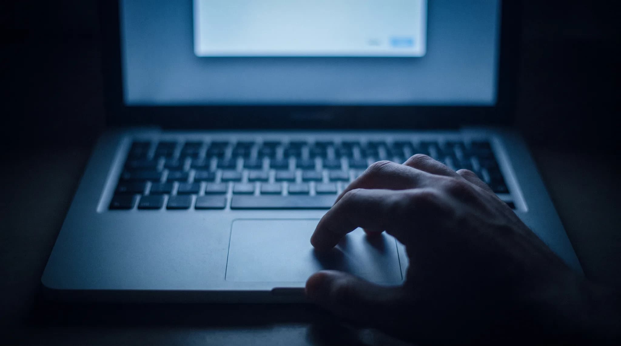Close cinematic still of a laptop keyboard and screen glow in a dark room, with a hand over the trackpad, suggesting quiet credential exposure.